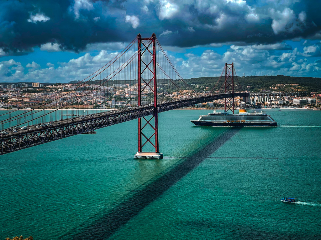 The 25th of April Bridge in Lisbon spans the Tagus River, with vehicles on it, a cruise ship below, and a small boat in the foreground, set against a cityscape and cloudy sky.